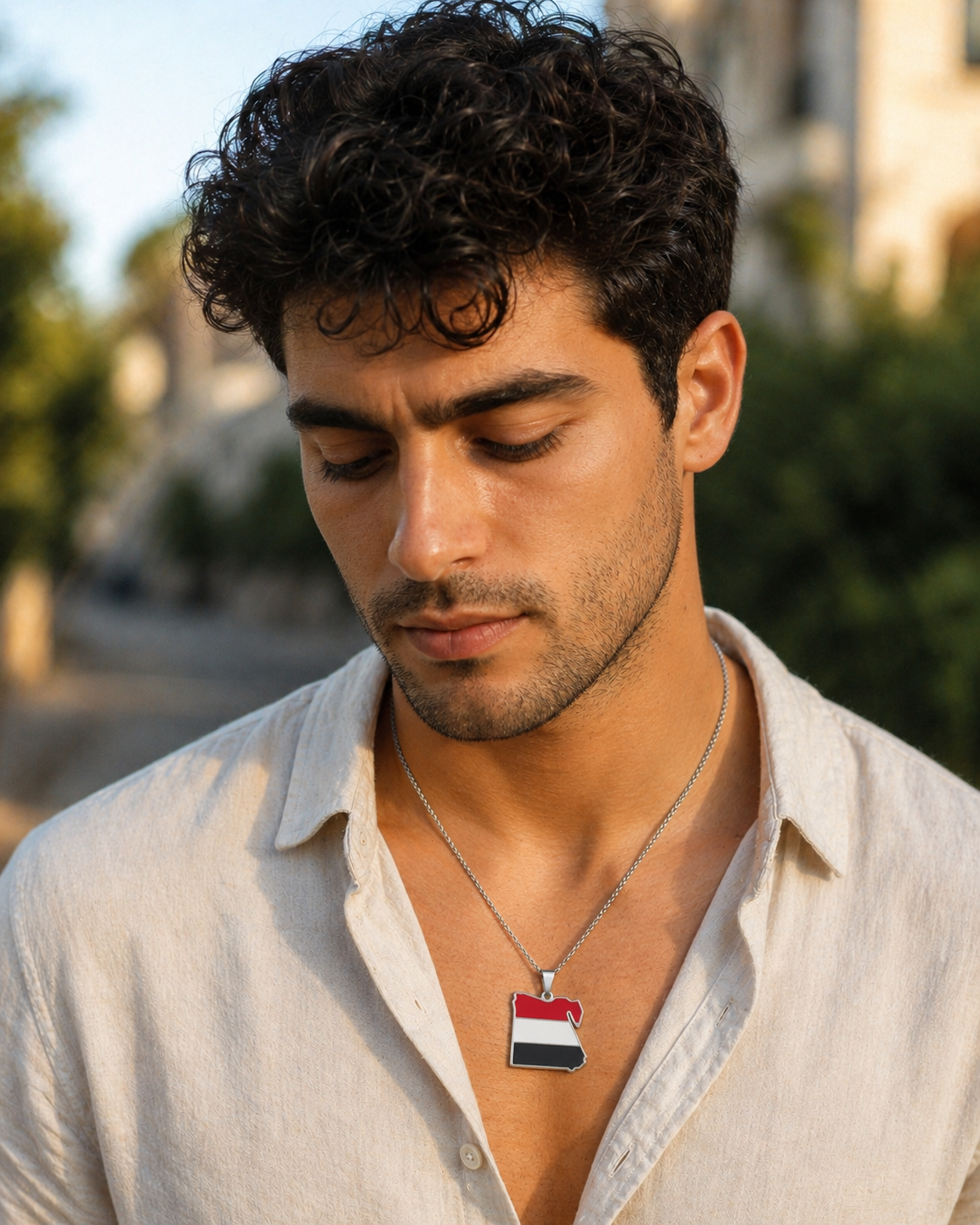 Silberne Halskette mit Ägypten-Karten-Anhänger in den Landesfarben Rot, Weiß und Schwarz – getragen von einem jungen Mann im beigen Leinenhemd, Outdoor-Porträt im warmen Sonnenlicht