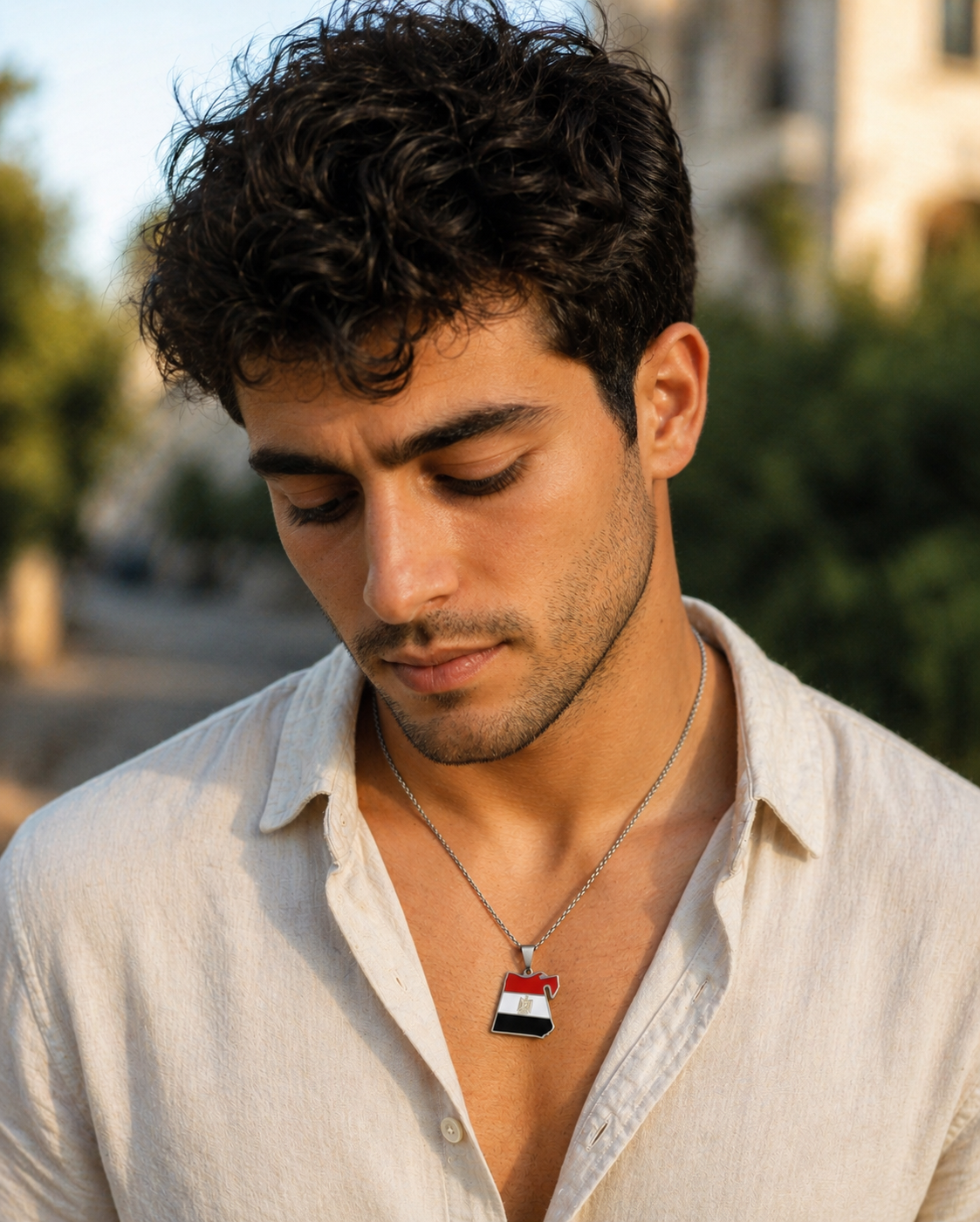 Silberne Halskette mit Ägypten-Karten-Anhänger in den Landesfarben Rot, Weiß und Schwarz mit goldenem Adler – getragen von einem jungen Mann im beigen Leinenhemd, Outdoor-Porträt im warmen Sonnenlicht