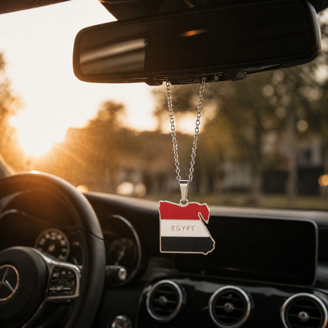 Silberner Auto-Rückspiegel-Anhänger mit Ägypten-Karten-Motiv in den Landesfarben Rot, Weiß und Schwarz mit Schriftzug "Egypt" – hängend im Mercedes-Innenraum bei Sonnenuntergang
