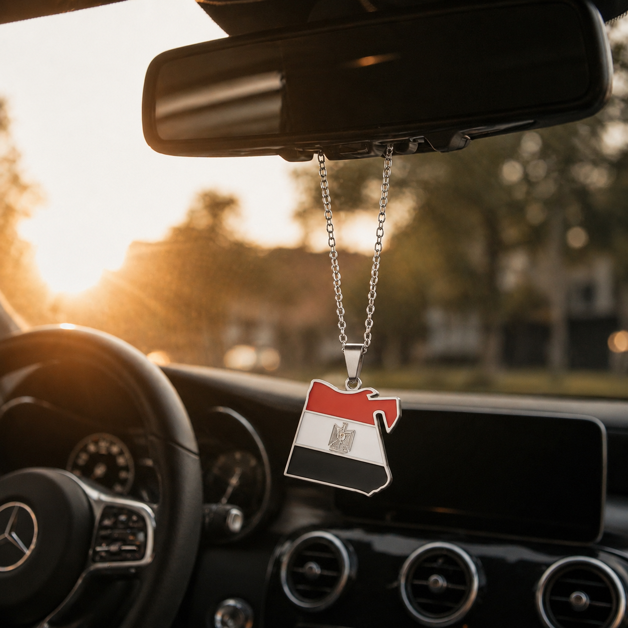 Silberner Auto-Rückspiegel-Anhänger mit Ägypten-Karten-Motiv in den Landesfarben Rot, Weiß und Schwarz mit goldenem Adler – hängend im Mercedes-Innenraum bei Sonnenuntergang