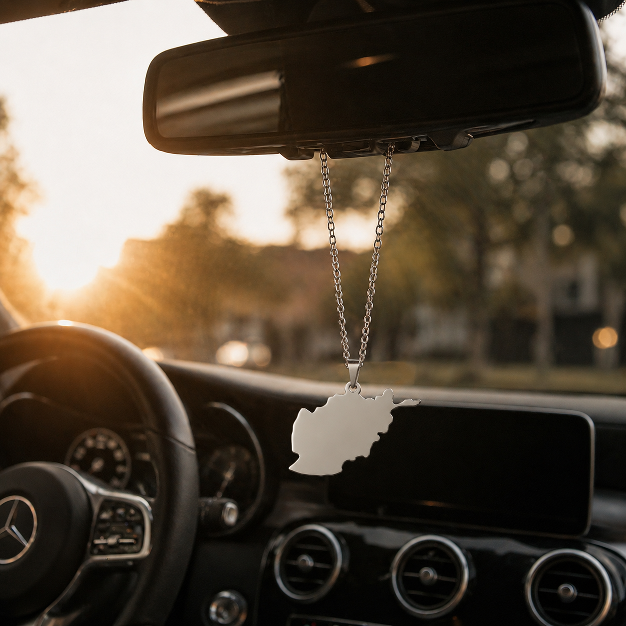 Silberner Auto-Rückspiegel-Anhänger mit mattiertem Afghanistan-Karten-Motiv ohne Farbfüllung – hängend im Mercedes-Innenraum bei Sonnenuntergang