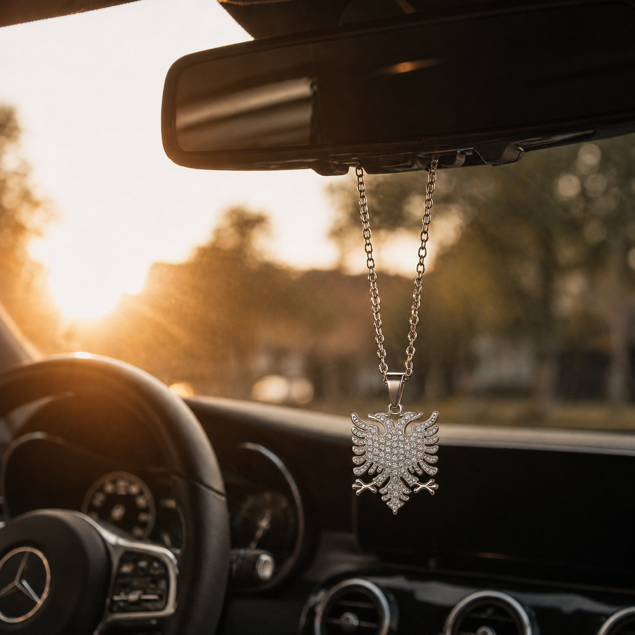 Silberner Auto-Rückspiegel-Anhänger mit albanischem Doppeladler-Motiv und Zirkonia-Steinen besetzt – hängend im Mercedes-Innenraum bei Sonnenuntergang