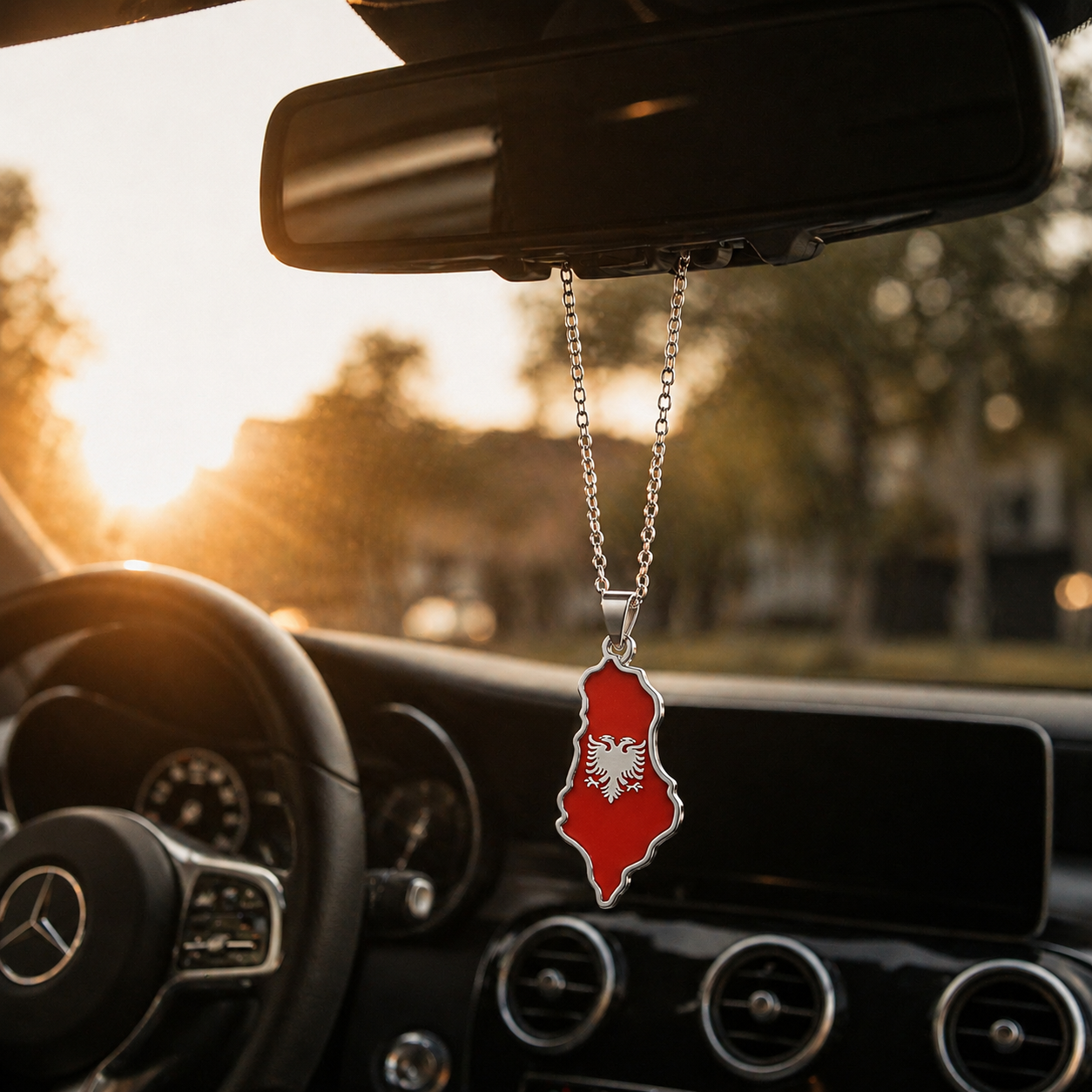 Silberner Auto-Rückspiegel-Anhänger mit Albanien-Karten-Motiv in Rot mit weißem Doppeladler – hängend im Mercedes-Innenraum bei Sonnenuntergang