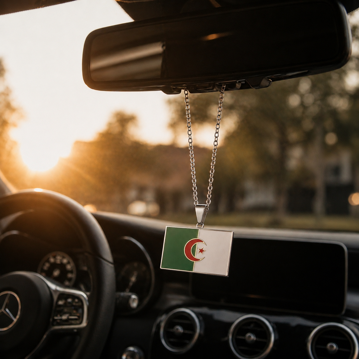 Silberner Auto-Rückspiegel-Anhänger mit rechteckigem Algerien-Flaggen-Motiv in den Landesfarben Grün und Weiß mit rotem Halbmond und Stern – hängend im Mercedes-Innenraum bei Sonnenuntergang