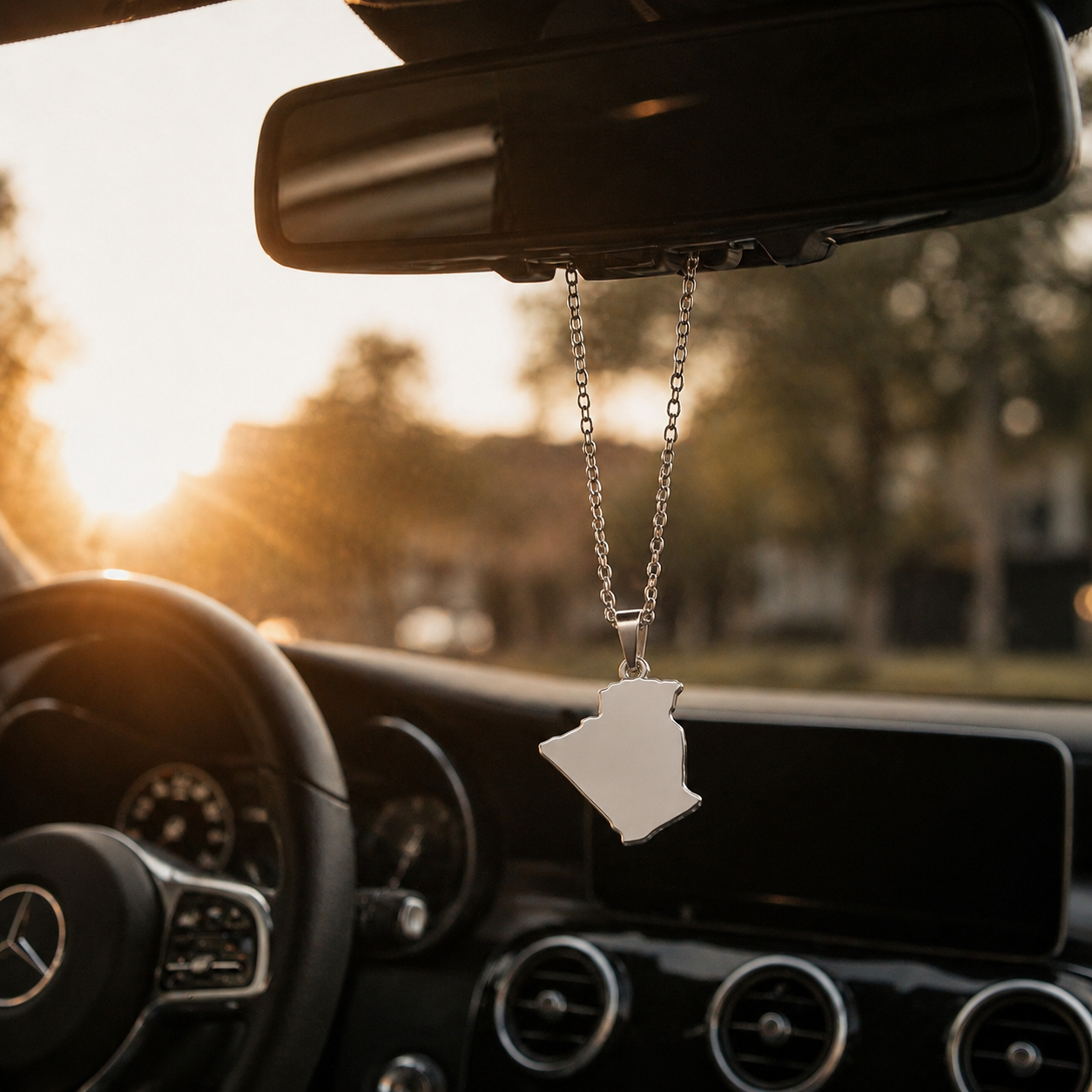 Silberner Auto-Rückspiegel-Anhänger mit mattiertem Algerien-Karten-Motiv ohne Farbfüllung – hängend im Mercedes-Innenraum bei Sonnenuntergang