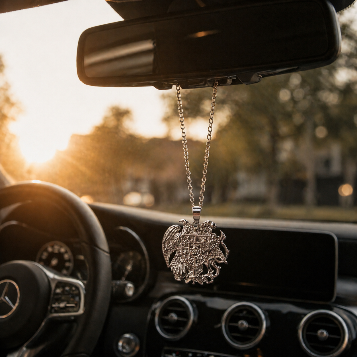 Silberner armenischer Wappen-Anhänger mit Adler und Schild, hängend am Rückspiegel eines Mercedes im warmen Sonnenuntergangslicht