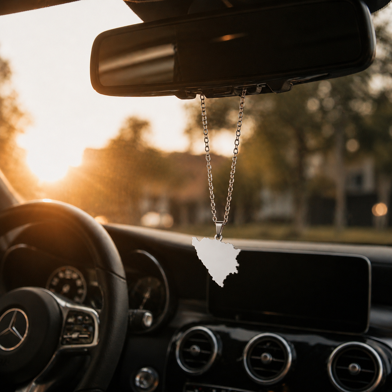 Silberner Auto-Rückspiegel-Anhänger mit mattiertem Bosnien-Karten-Motiv ohne Farbfüllung – hängend im Mercedes-Innenraum bei Sonnenuntergang