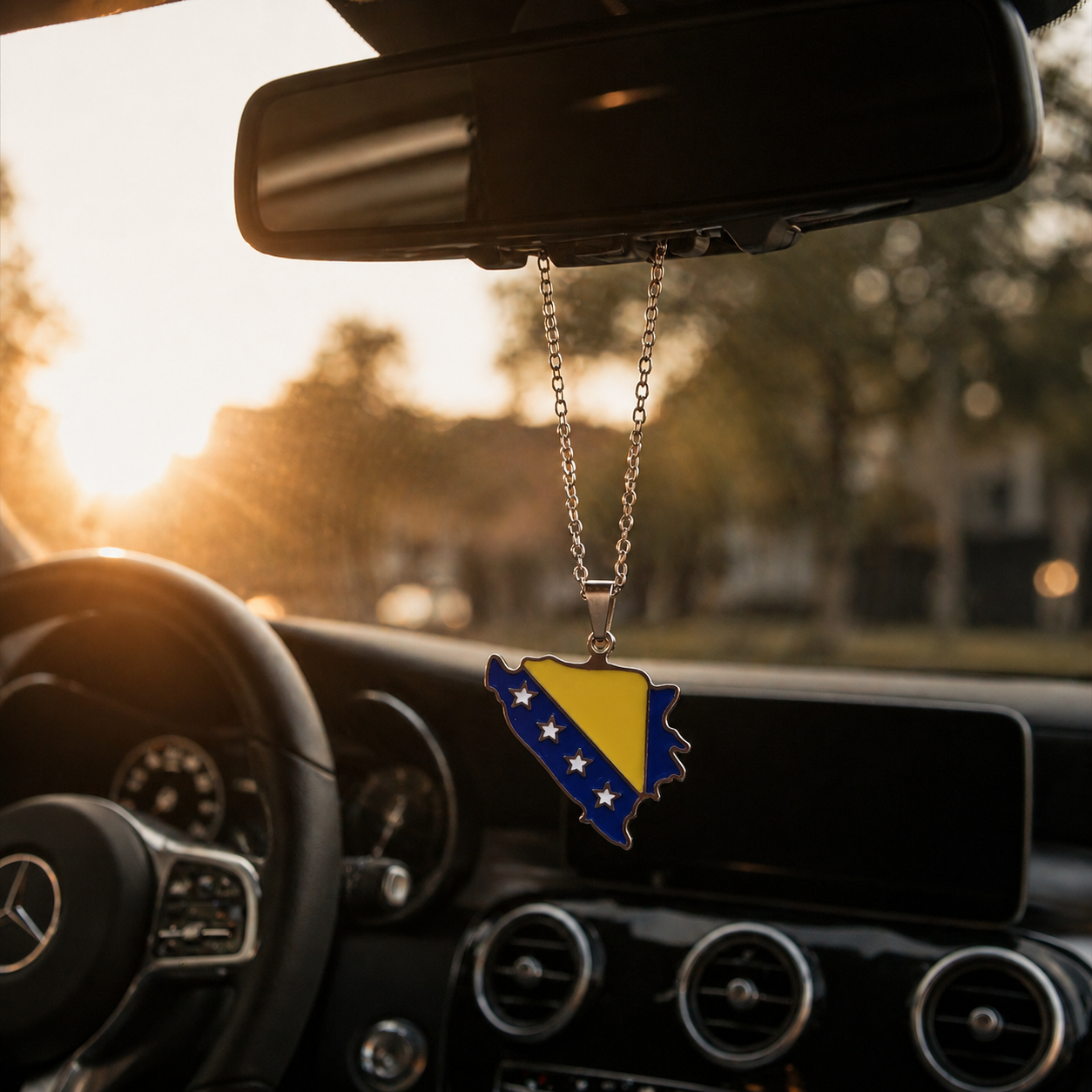 Silberner Auto-Rückspiegel-Anhänger mit Bosnien-Karten-Motiv in den Landesfarben Blau und Gelb mit weißen Sternen – hängend im Mercedes-Innenraum bei Sonnenuntergang