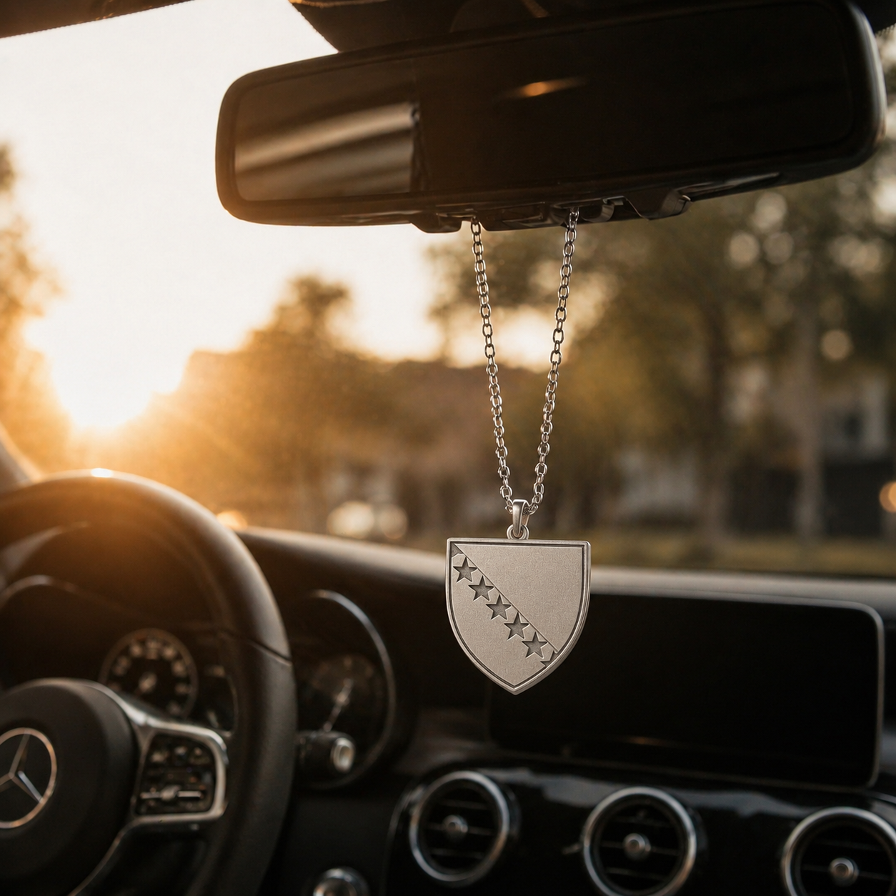 Silberner Auto-Rückspiegel-Anhänger mit Bosnien-Wappen in Schildform und diagonaler Sternenreihe – hängend im Mercedes-Innenraum bei Sonnenuntergang