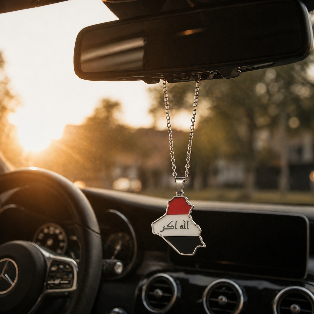 Silberner Auto-Rückspiegel-Anhänger mit Irak-Karten-Motiv in den Landesfarben Rot, Weiß und Schwarz mit arabischer Schrift – hängend im Mercedes-Innenraum bei Sonnenuntergang