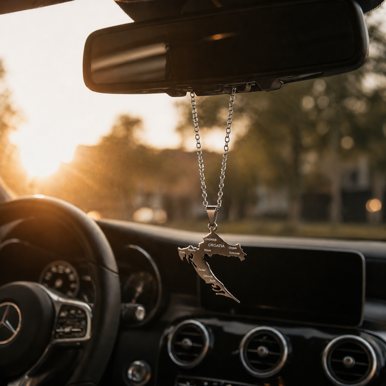 Silberner Kroatien-Karten-Anhänger mit gravierten Städtenamen hängend am Rückspiegel eines Mercedes im warmen Sonnenuntergangslicht