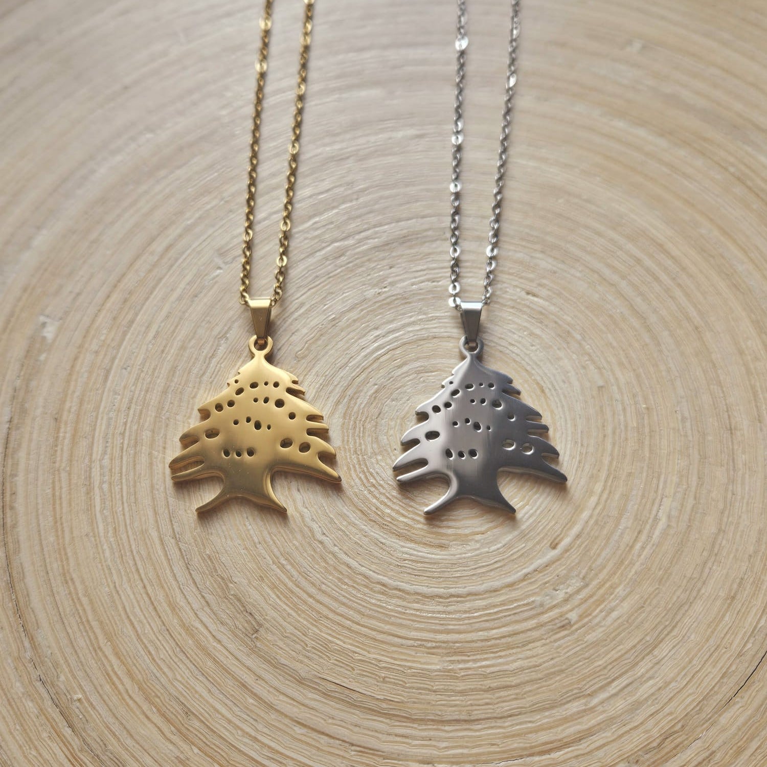 Zwei Halsketten mit libanesischer Zeder-Anhänger in Gold und Silber, nebeneinander auf einer Holzscheibe liegend