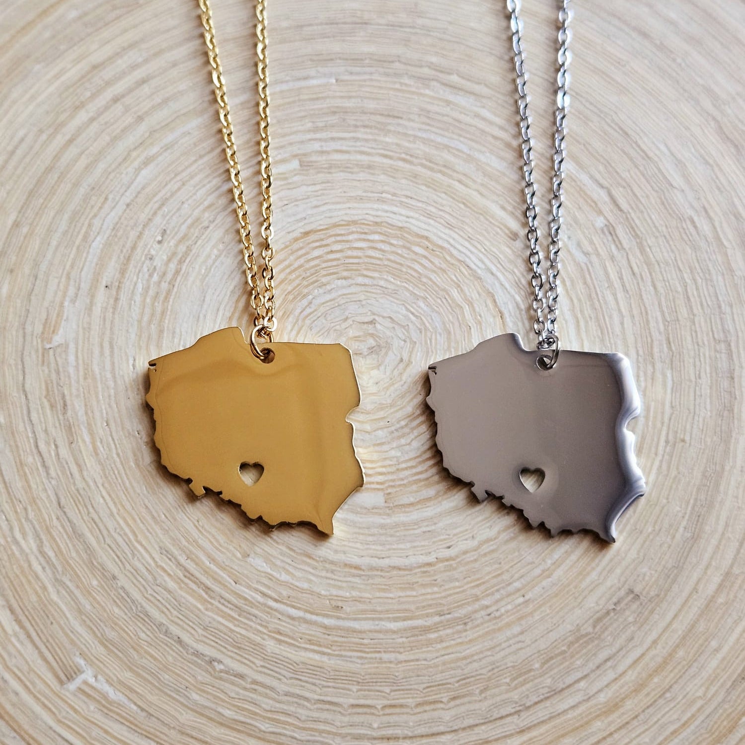 Zwei Halsketten mit massivem Polen-Karten-Anhänger mit ausgestanztem Herz – in Gold und Silber – nebeneinander auf einer Holzscheibe liegend