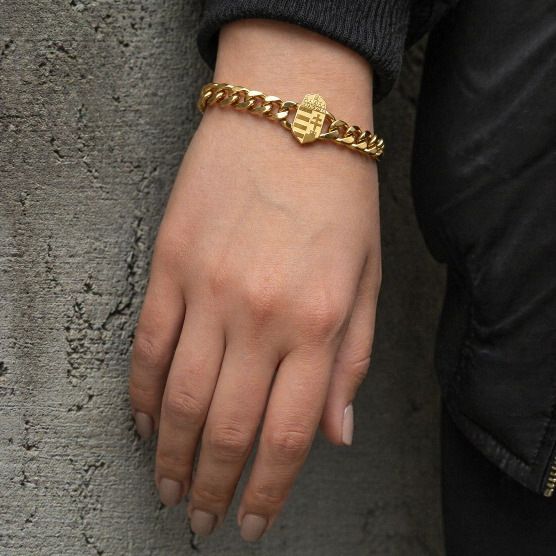 Goldenes Panzerarmband mit ungarischem Wappen-Element, getragen an einem weiblichen Handgelenk vor grauer Betonwand